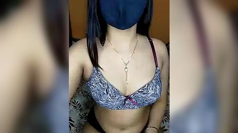 Media: Video of a person with a blue mask, wearing a blue and white zebra-patterned bra with a pink bow, and a gold necklace. The background is blurred, suggesting a focus on the subject.