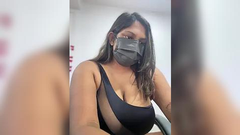 Media: Video of a woman with medium skin tone, wearing a black face mask and a revealing black one-shoulder top, seated indoors, blurred background.