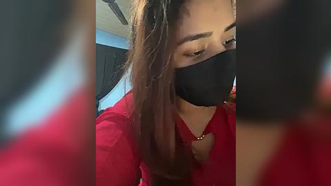 Media: A video of a woman with long brown hair, wearing a black face mask, red top, and heart necklace, sitting indoors.