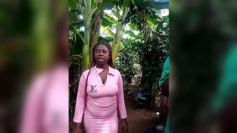 Media: Video of a Black woman with braided hair in a light pink V-neck shirt standing in a lush, green, tropical garden with banana leaves and a green tarpaulin overhead.