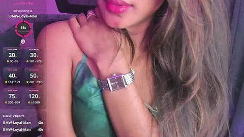 Media: Video of a light-skinned woman with long, wavy brown hair, wearing a green strapless top, and a silver necklace, partially visible. Background is dark and blurry.