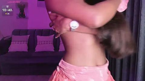 Media: Video of a woman in a pink satin top and skirt, holding her bra, in a dimly lit living room with a dark couch and decorative pillows.