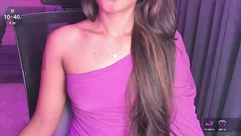 Media: A video of a woman with long, wavy brown hair, wearing a one-shoulder purple dress, sitting in a dimly lit room with purple lighting.