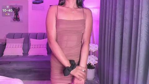 Media: A video of a slender woman with fair skin and long dark hair, wearing a tight, beige, spaghetti-strap dress, standing in a dimly lit, modern bedroom with purple lighting and white flowers.