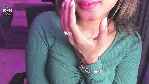 Media: Video of a woman with light brown skin, medium breasts, and a green ribbed sweater. She has a silver nose ring and is resting her face in her hand. Background shows a purple room with a couch.