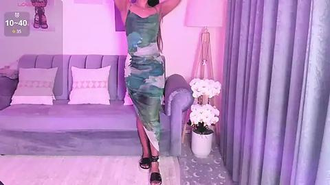 Media: Video of a woman in a green, white, and black abstract dress, posing on a light gray couch with white pillows, surrounded by lavender curtains and a floral arrangement.