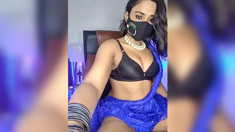 Media: Video of a woman with dark skin, wearing a black bra, blue lace dress, gold jewelry, and a black mask, seated indoors with blurred background.