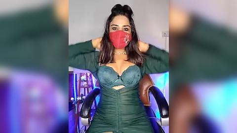 Media: Video of a South Asian woman with medium brown skin, wearing a red mask, dark green dress, and brown hair tied up, seated in a chair, indoors with blurred background.