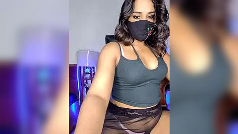 Media: Video of a young woman with medium brown skin, wearing a black face mask, a grey crop top, and black panties, sitting in a dimly lit room with a blue light, blurred background.