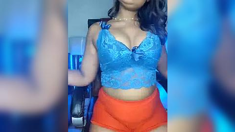 Media: Video of a woman with medium brown skin, wearing a blue lace bralette and orange shorts, sitting in a gaming chair with a blurred background.