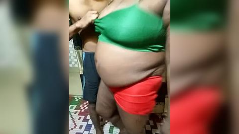 Media: Video of a pregnant woman with fair skin, wearing a green sports bra and red shorts, standing in a bathroom with a tiled floor.