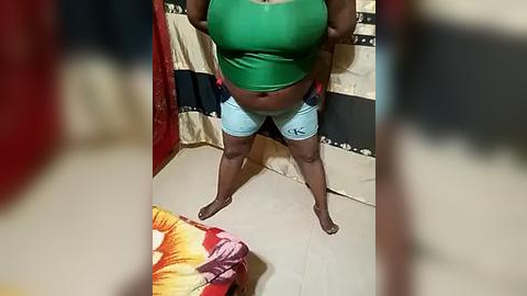 Media: Video of a pregnant woman in a green tank top and light blue shorts, standing barefoot in a room with beige walls and a colorful blanket on the bed.