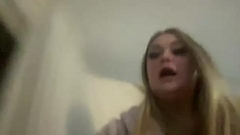 Media: Video of a distressed woman with long blonde hair, wearing a beige top, mouth open, eyes wide, blurry background, suggesting a tense, possibly emotional moment.