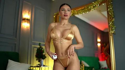 Media: Video of a fit, tan-skinned woman with dark hair in a sleek updo, wearing gold lingerie, holding a cigarette, standing in a luxurious, dimly-lit room with gold-framed mirror and green sofa.