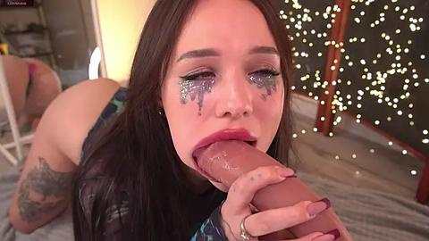 Media: Video of a young woman with long dark hair and fair skin, wearing glittery eye makeup, performing oral sex on a large, circumcised penis in a dimly lit room with string lights and a white chair in the background.