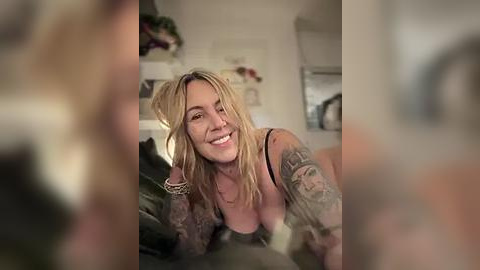 Media: Video of a smiling, blonde, tattooed woman with medium breasts, wearing a black bra, leaning on a bed. Background shows a blurred, softly lit room with plants and furniture.