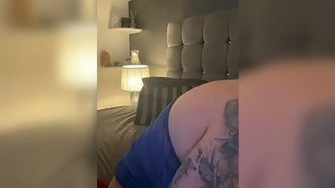 Media: Video of a dimly lit bedroom showing a person's tattooed back and a bed with a grey headboard, a lamp, and a side table.