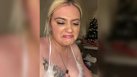 Media: Video of a middle-aged woman with fair skin and blonde hair, wearing a white feathered top, with a visible tattoo on her chest, looking down, in a dimly lit room with a Christmas tree in the background.