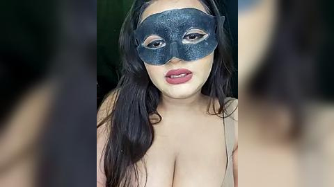 Media: Video of a woman with long, dark hair, wearing a dark blue glittery masquerade mask, revealing full lips and cleavage. The background is blurred and dark.