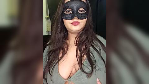 Media: Video of a woman with long brown hair, wearing a black lace mask and a green knit top, with a blurred background.