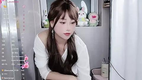 Media: A video of an Asian woman with long, dark hair and fair skin, wearing a white blouse, sitting in a room with a pink bunny plush toy on a shelf.