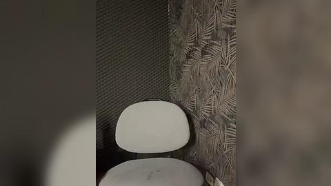 Media: Video of a modern, white, molded plastic chair with a curved backrest, positioned against a textured, dark green wall with a pattern of leafy vines. The background is out of focus, emphasizing the chair's minimalistic design.