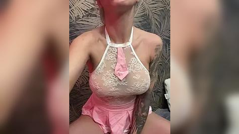 Media: Video of a woman in a pink lace bodysuit with a white halter neck and a pink tie, sitting on a bed with a fern-patterned headboard. She has a tattoo on her left arm.