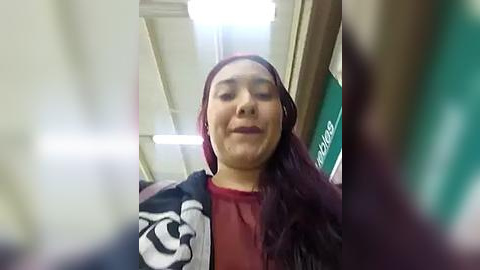 Media: Video of a young woman with long dark hair, wearing a red hijab and a black jacket with a white design, standing in a brightly lit hallway with fluorescent lights.