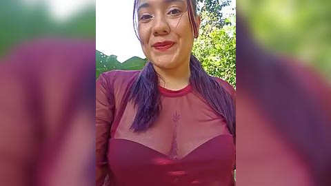 Media: A video of an Asian woman with light skin, long purple hair in pigtails, wearing a sheer, maroon, long-sleeved top that reveals her cleavage. She's smiling outdoors with green foliage in the background.