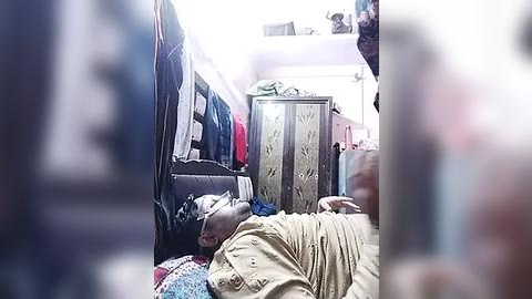 Media: Video of a cramped, dimly lit room with a man lying on a bed, wearing a beige robe. The background includes a wardrobe and colorful clothing. The scene appears cramped and somewhat disorganized.