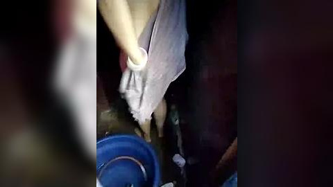 Media: A video captures a person in a sleeveless shirt and apron cleaning a dark, possibly wooden surface with a white cloth. A blue bucket and a green bottle are visible in the background. The image is dimly lit.