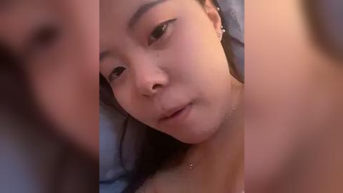 Media: A close-up video of a young Asian woman with light skin, dark hair, and small earrings, lying on a bed. Her expression is calm. The background is blurred.