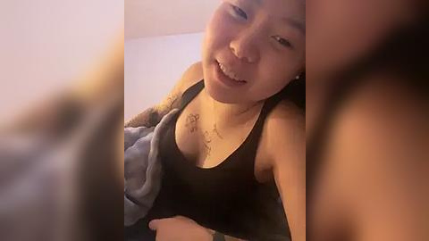 Media: Video of a smiling East Asian woman with light skin, wearing a black tank top, lying on a gray blanket. She has tattoos on her chest and arm.