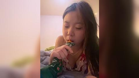 Media: A candid video captures an Asian woman with long brown hair, wearing a colorful floral top, eating a green snack bag while lying on a bed with a beige wall in the background.