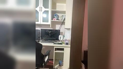 Media: Video of a cluttered, beige-colored home office with a white desk, black chair, monitor, keyboard, and various office supplies. Shelves hold books and decorative items. Background includes a blurred person.