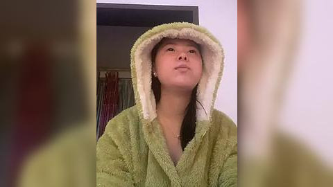 Media: A video of a young Asian woman in a lime-green, fuzzy robe with a hood, standing indoors with a neutral expression. The background features a partially open door and colorful, vertical curtains.