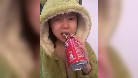 Media: Video of an Asian baby in a green, plush dinosaur costume, drinking from a red can of soda. The background is blurry.