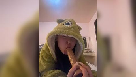 Media: Video of a young child in a green dinosaur pajama costume with a large, round blue eye on the hood, licking a long, brown lollipop. Background shows a dimly lit room with a white dresser and a mirror.