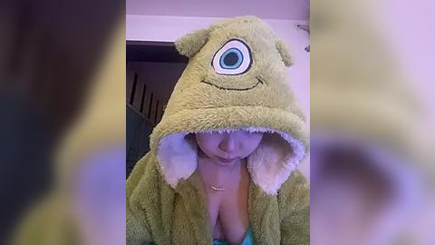 Media: Video of a woman wearing a green, plush monster hoodie with large eyes, covering her chest, indoors with blurred background.