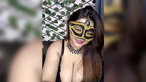 Media: Video of a smiling woman with long brown hair, wearing a revealing black and gold masquerade mask and a black lace bra, against a blurred green and white patterned backdrop.