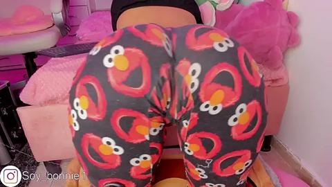 Media: Video of a person wearing Elmo-patterned leggings, bent over a pink bed with a plush toy, in a pink room.