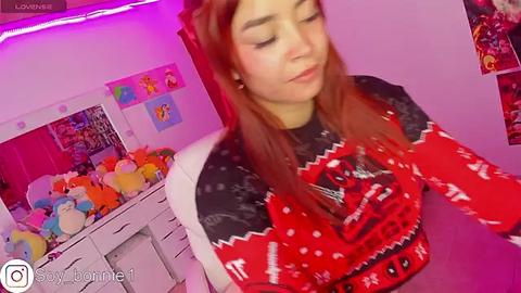 Media: Video of a young woman with light skin and long red hair, wearing a festive red and black sweater, surrounded by stuffed animals and colorful posters in a cozy room.