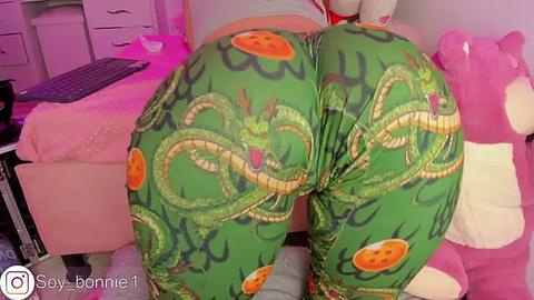 Media: Video of a person with light skin, wearing green leggings adorned with cartoonish dragon patterns and a white top, bending over in a cozy room filled with plush toys and a pink blanket.