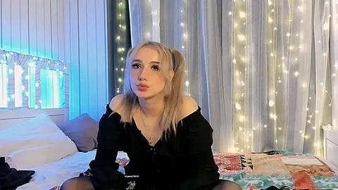 Media: Video of a blonde, fair-skinned woman with pigtails, wearing a black off-shoulder top, sitting on a bed with a multicolored quilt, surrounded by soft, glowing fairy lights and sheer curtains.