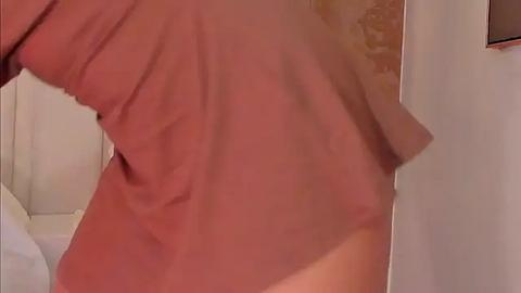 Media: A close-up video of a person's lower back, wearing a pink shirt, standing in a bathroom with beige tiles. The image is slightly blurry.