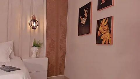 Media: A minimalist bedroom with a modern design, featuring a white bed, beige walls, a brown-framed floral print, and a hanging pendant light with a metallic texture.