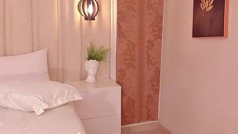 Media: Video of a minimalist bedroom with beige walls, white bedding, a white side table, a potted green plant, a decorative urn, and a brown damask curtain.