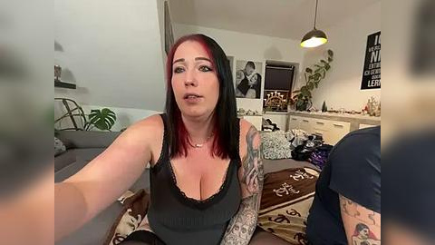 Media: Video of a pale-skinned woman with red and black hair, wearing a black lace-trimmed tank top, seated on a beige couch in a dimly lit living room with potted plants, posters, and a hanging light.