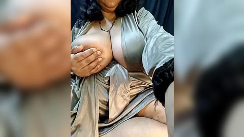 Media: Video of a voluptuous, dark-skinned woman with curly hair, wearing a silky, metallic silver robe, covering her ample breasts with her hands. Background is blurred, focusing attention on her figure.