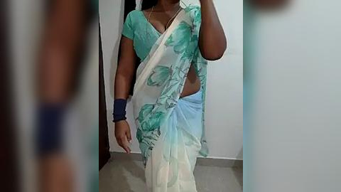 Media: Video of a woman in a turquoise and white saree with a floral pattern, wearing a dark blue bracelet, taking a selfie in a dimly lit room with a white wall and tiled floor.